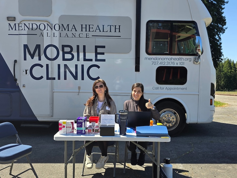 2 women give the thumbs up outfront of the Mendonoma Health Alliance Mobile Health Bus