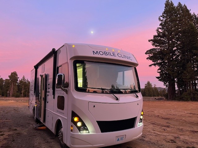 A white mobile clinic vehicle parked in an open, rural area at sunset, with a pink and purple sky in the background. Tall pine trees stand nearby, and the words “MOBILE CLINIC” are printed above the vehicle’s windshield. The clinic lights are on, and the side door of the vehicle appears open, suggesting it is ready to provide medical services.
