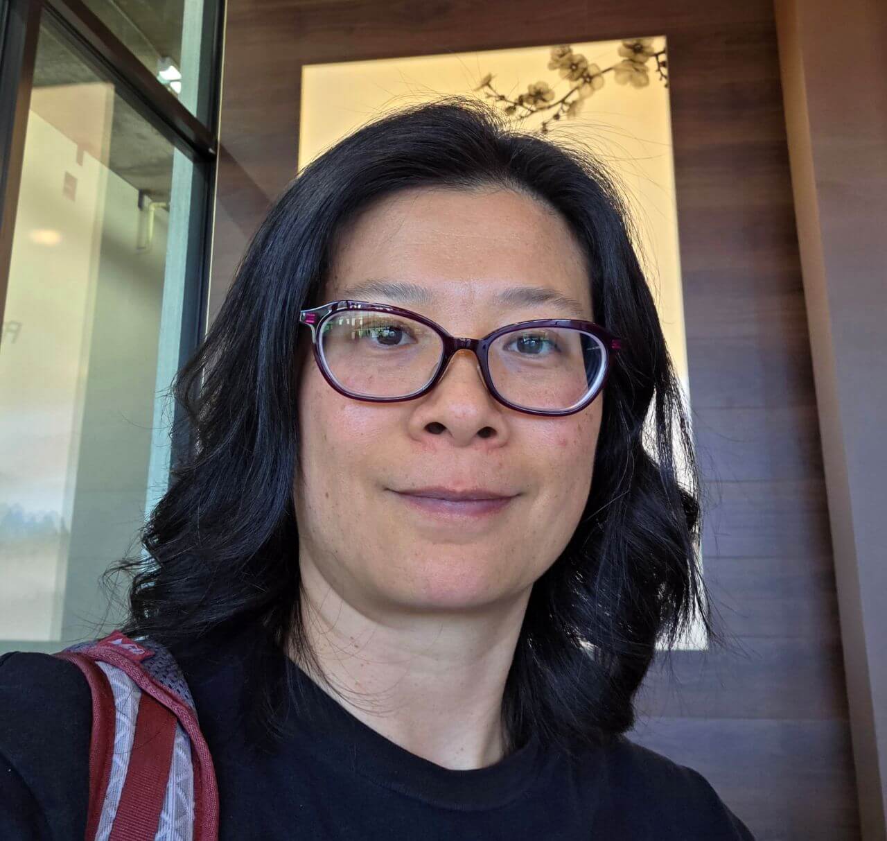 Head-and-shoulders portrait of a woman with shoulder-length dark brown hair wearing burgundy-framed glasses and a black top. She is looking at the camera with a calm expression. The background includes warm wood paneling and a softly lit cream-colored wall, with a hint of a red strap over her shoulder.