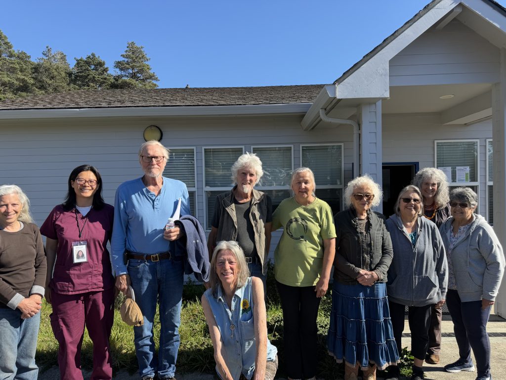 A group of about ten older adults and one middle-aged adult stand and sit together outside a light-gray building on a sunny day. Most are smiling and facing the camera. One person sits in front while the others stand behind in a line. They wear casual clothing in a variety of colors, including blue, green, and maroon. The building has windows and a covered entryway, and trees and a clear blue sky are visible in the background.