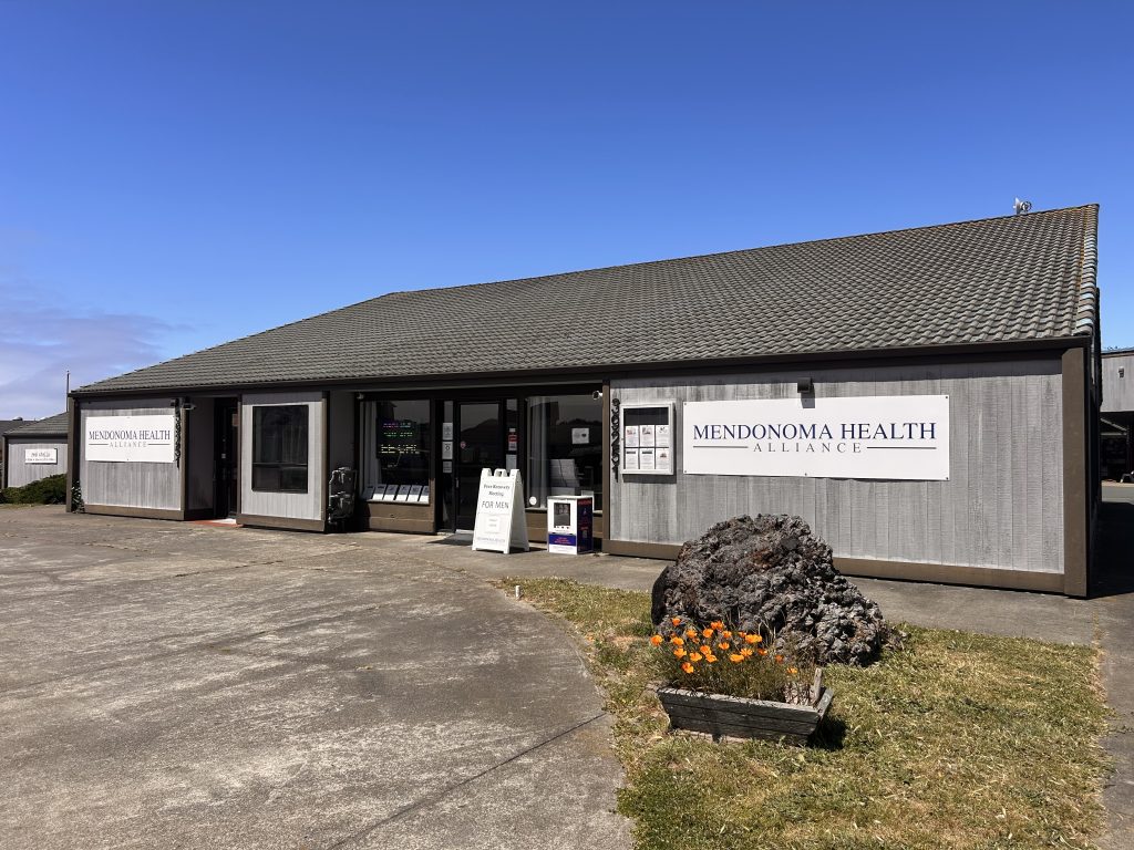 Exterior view of the Mendonoma Health Alliance building on a sunny day, featuring gray siding, large white signs with the organization’s name, glass front doors, and a paved driveway in front. A large decorative rock and planter with orange flowers sit near the entrance under a clear blue sky.